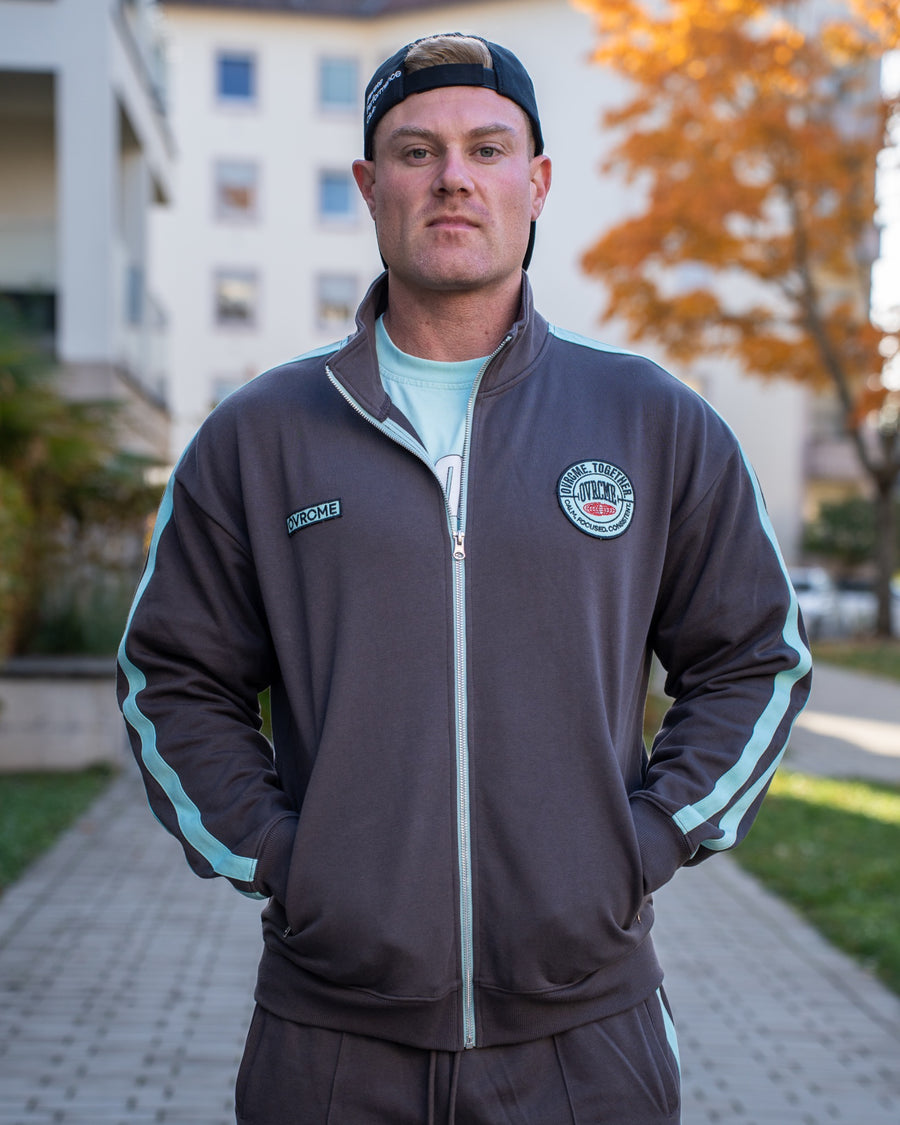 Vorderansicht der OVRCME CLUB Track Jacket in Anthrazit/Mint mit Stehkragen, Reißverschluss und Brustpatches.